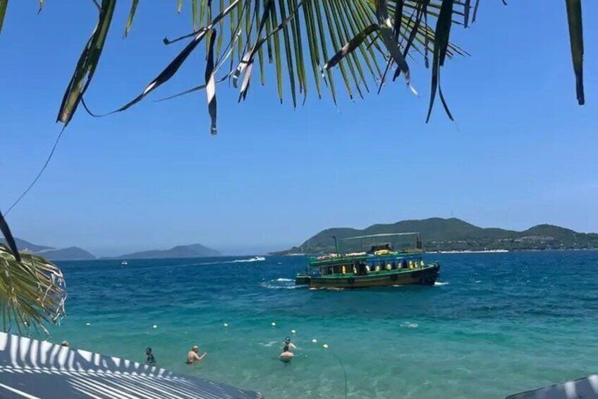 A boat on a clear blue sea with people swimming, palm trees, and distant hills. 2