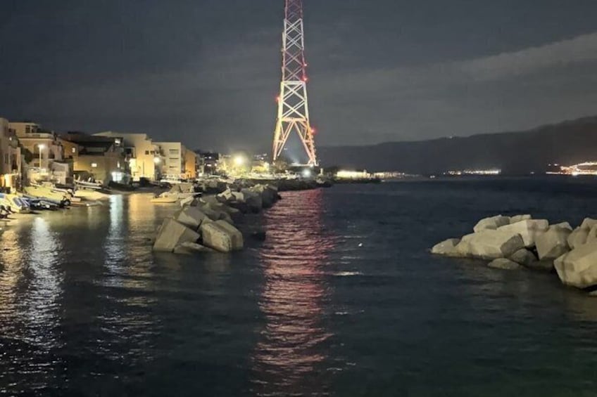A nighttime coastal scene with a lit tower, calm water, and buildings along the shore. 5