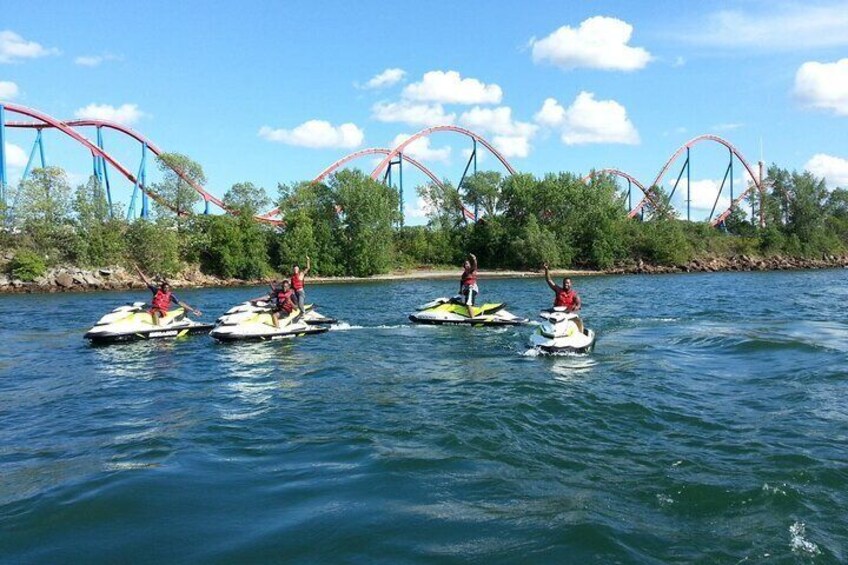 Four people jet-skiing on a lake with roller coasters in the background. 4