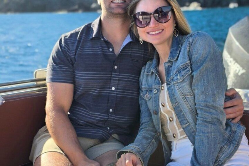 Two people on a boat with water and a rocky shoreline in the background. 4
