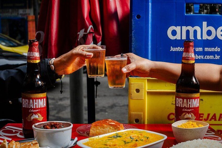 Two people toasting with drinks, a bottle of Brahma beer, a bowl of food, and a yellow box with text. 3