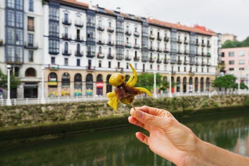A hand holding a yellow flower with a brown center, a river, and buildings with balconies in the background. 1