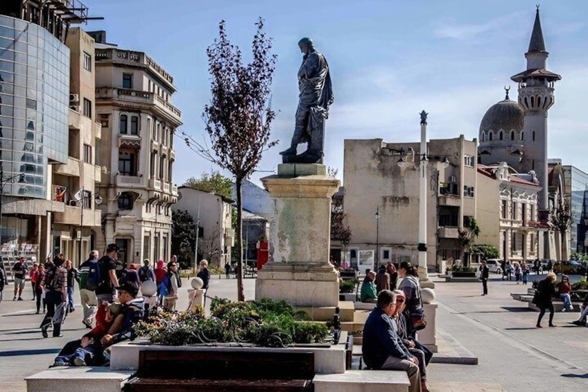 A public square with a statue, people walking, and a mosque in the background. 4