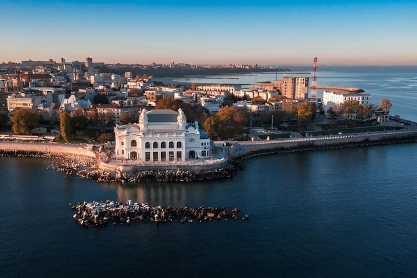 A coastal cityscape with a prominent white building, a lighthouse, and a dense urban area in the background. 1