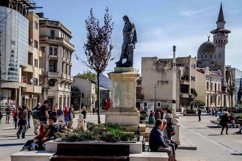 A public square with a statue, people walking, and a mosque in the background. 3