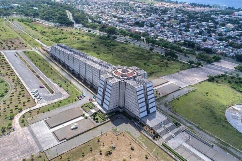 A large modern building with a unique rooftop structure, surrounded by green spaces and a cityscape in the background. 1