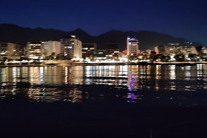 A nighttime cityscape with buildings reflected in calm water, a mountain in the background, and a clear sky. 3