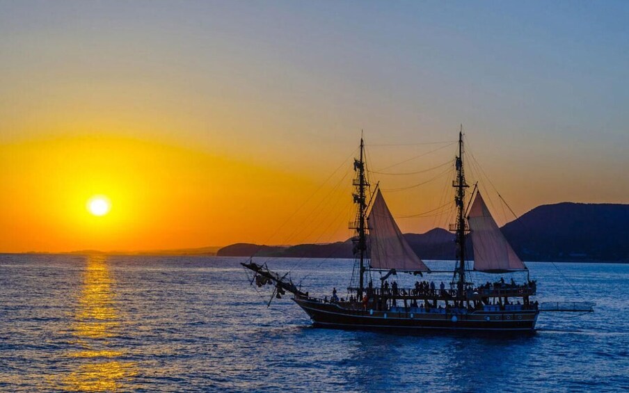 A sailing boat with multiple masts and sails, passengers, and a sunset over water with distant land. 1