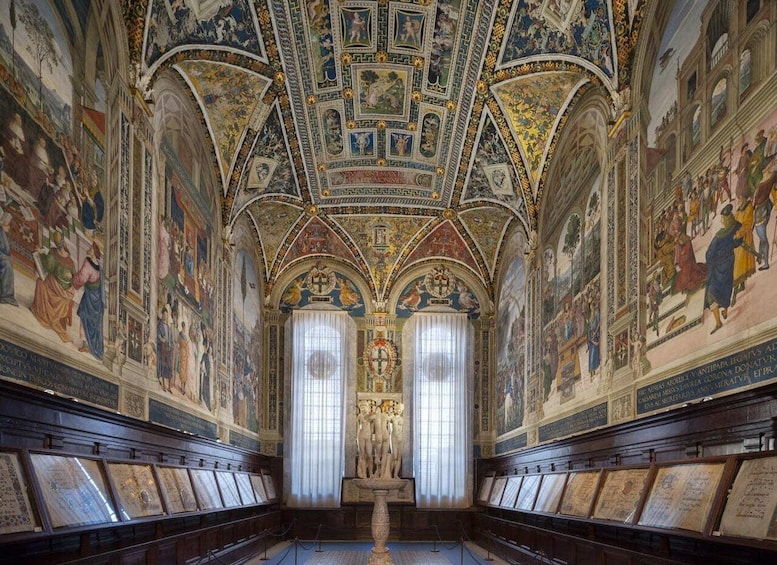 An ornate hall with frescoes, a central statue, and framed documents on the walls. 4