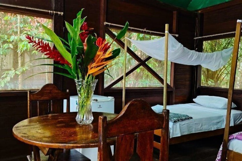 A wooden room with a bed, a table with a vase of flowers, and a window with a view of greenery. 3