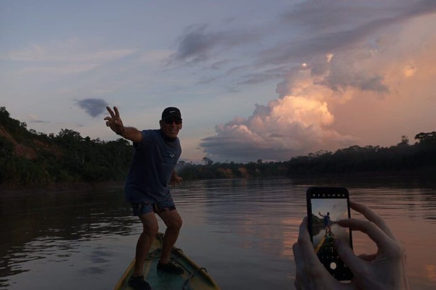 A person standing on a boat, another person taking a photo, and a sunset over water. 3