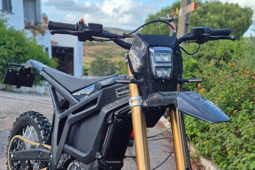 A black dirt bike parked on a driveway with a house and greenery in the background. 4