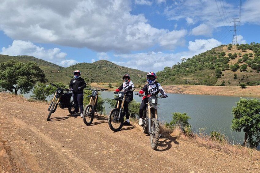 Three mountain bikers on a dirt path near a lake with hills in the background. 1