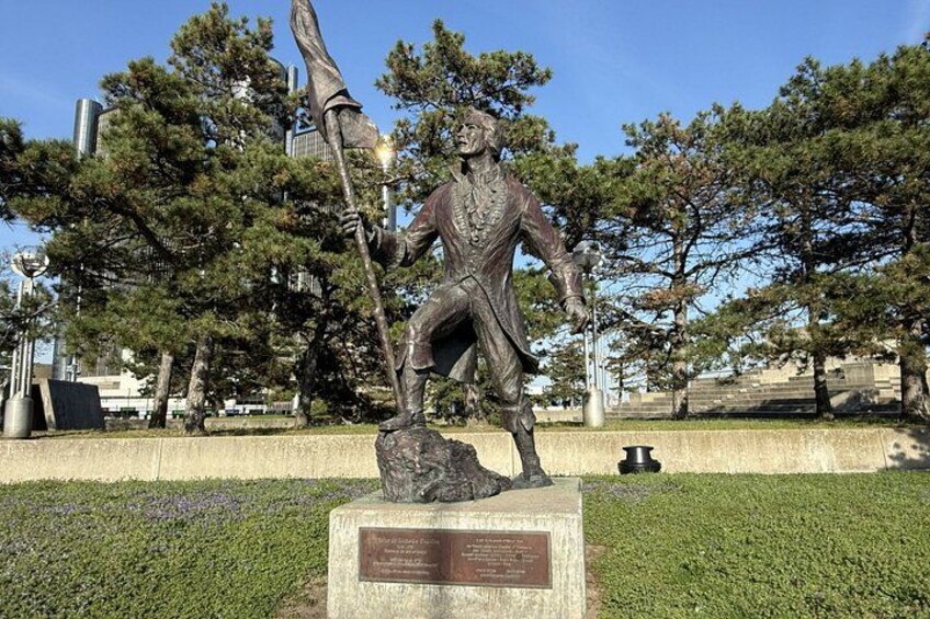 A bronze statue of a man holding a staff in a park with trees and a plaque. 3