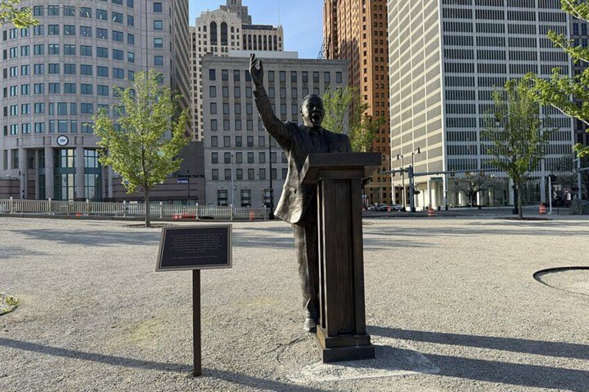 A bronze statue of a man with raised arms, standing on a pedestal in an urban square with modern buildings in the background. 4