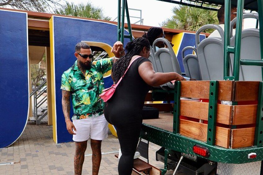 A man in a green shirt and white shorts, a woman in a black outfit, and a green utility vehicle with stacked wooden crates. 3
