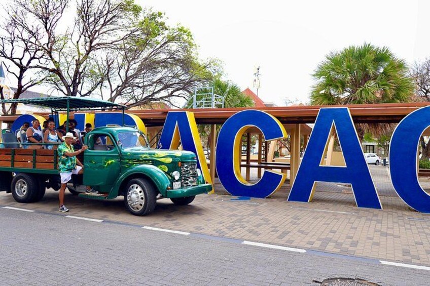 A green vintage truck parked in front of a large blue and yellow sign with the letters 'CACAO' on a paved area with trees and a building in the background. 1