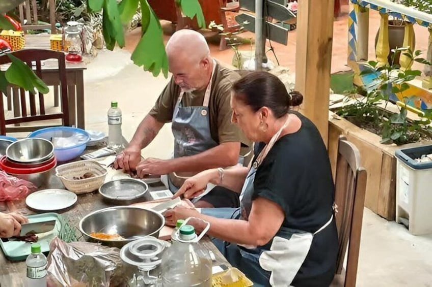 Two people preparing food at an outdoor table with various kitchen utensils and ingredients. 1