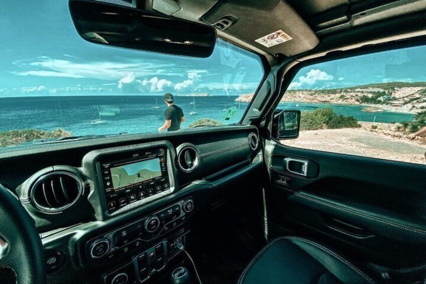 A car interior with a view of the ocean and a person standing by the shore. 1