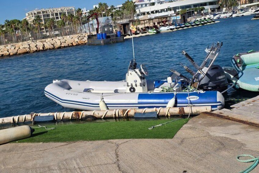 A white and blue inflatable boat docked at a marina with a rocky shoreline and palm trees in the background. 2