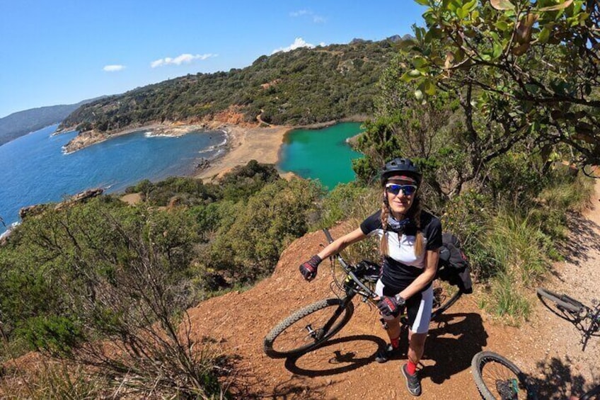A mountain biker on a dirt path with a scenic view of a turquoise lake and coastal landscape. 1