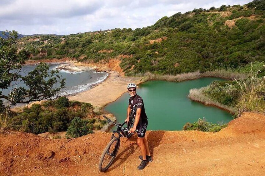 A person standing on a dirt path with a mountain bike, overlooking a green lake and a beach. 3