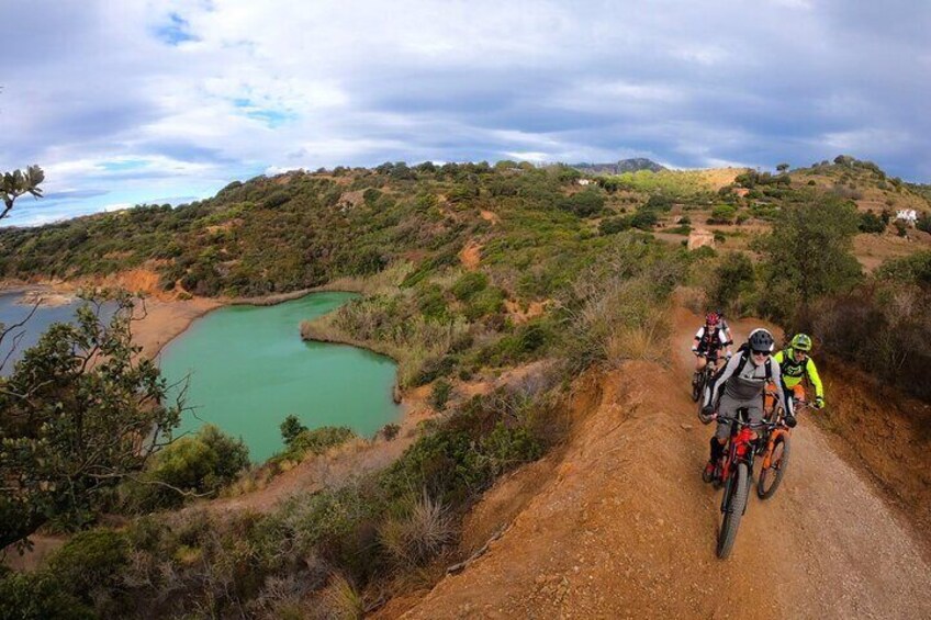 A dirt road with cyclists, a green lake, and hilly terrain. 2