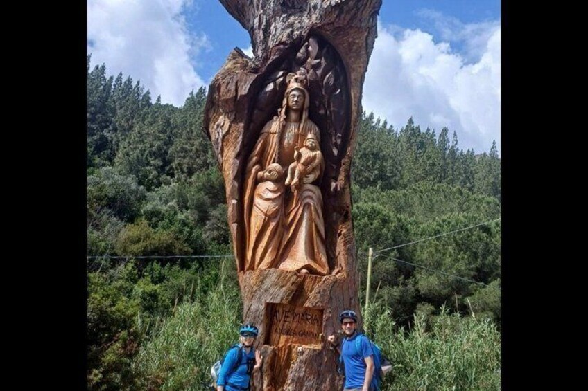 A large wooden carving of a seated figure with a child, set in a natural landscape with trees and a clear sky. 2