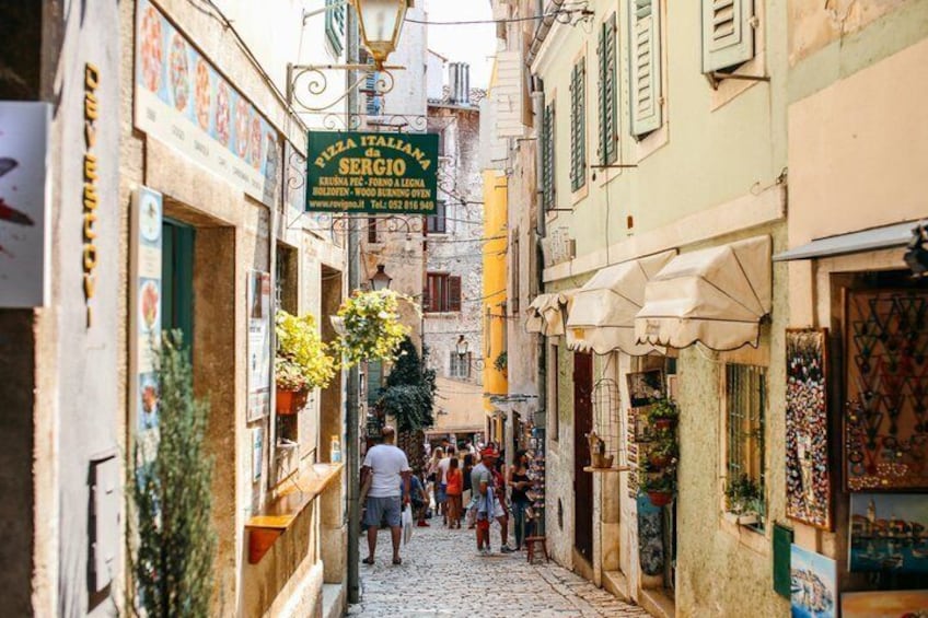 A narrow street with shops, a sign for 'PIZZA ITALIANA SERGIO,' and people walking. 4