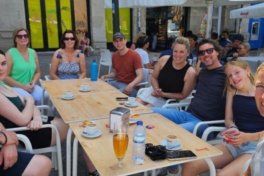 A group of people sitting at a wooden table outdoors, with drinks and a coffee machine on the table. 3