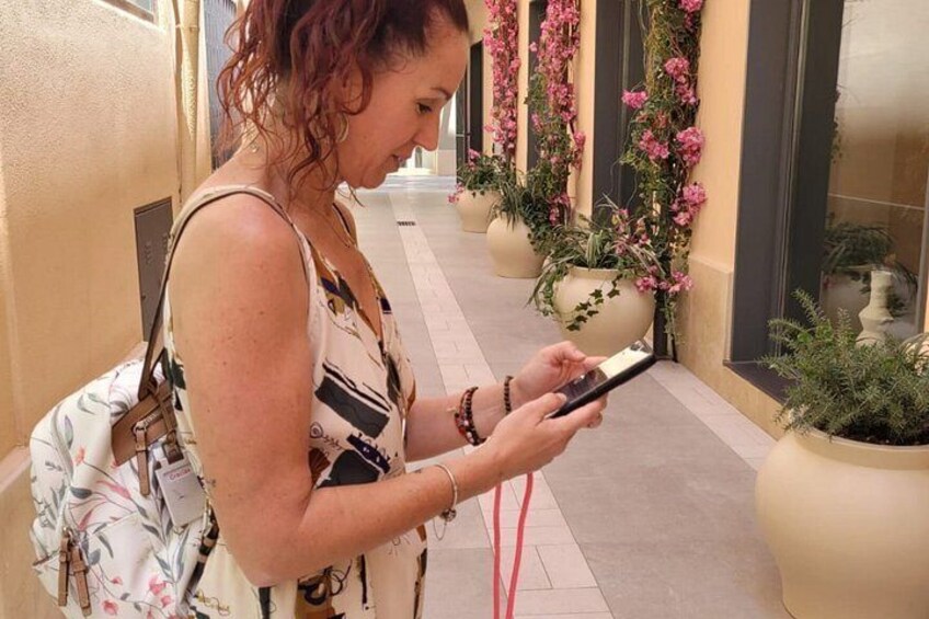 A person standing on a sidewalk, holding a smartphone, with potted plants and a building in the background. 3