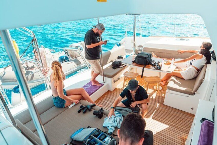 A group of people on a boat with a person standing and using a mobile device, another person sitting on the deck, and a third person sitting on a cushion. 5