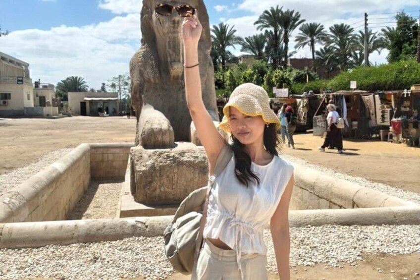 A person standing in front of a large stone structure with a raised arm, a sandy area, and a market in the background. 5