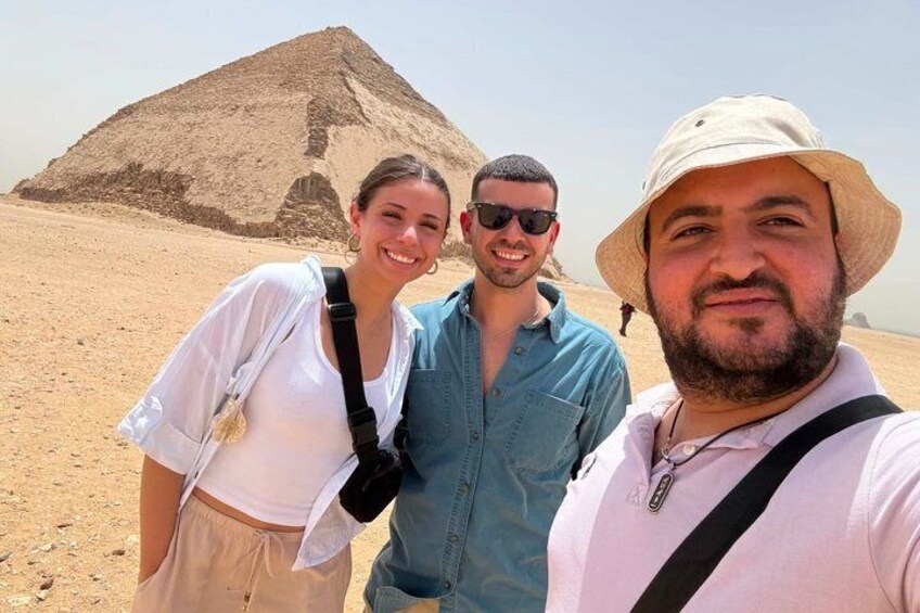 Three people standing in front of a pyramid in a desert. 1