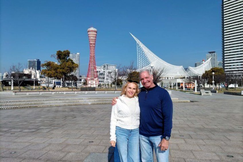 A couple standing in a plaza with a red tower and a white building in the background. 1