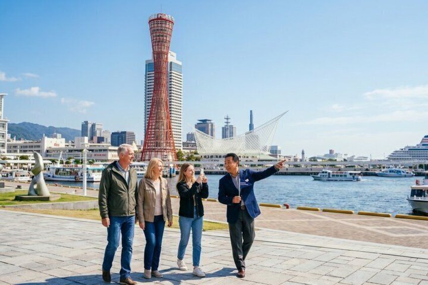 A group of people standing by a waterfront with a distinctive red tower and a white sail-like structure in the background. 1
