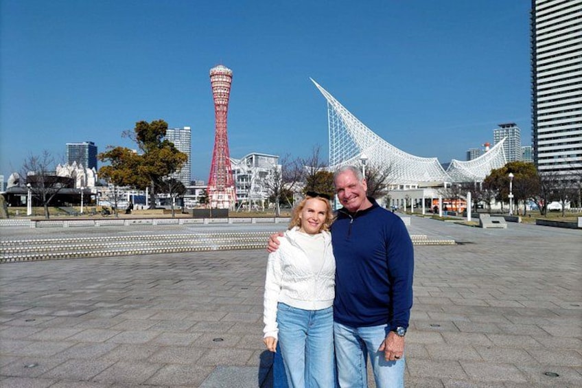 A couple posing in a plaza with a red tower and a white building in the background. 1