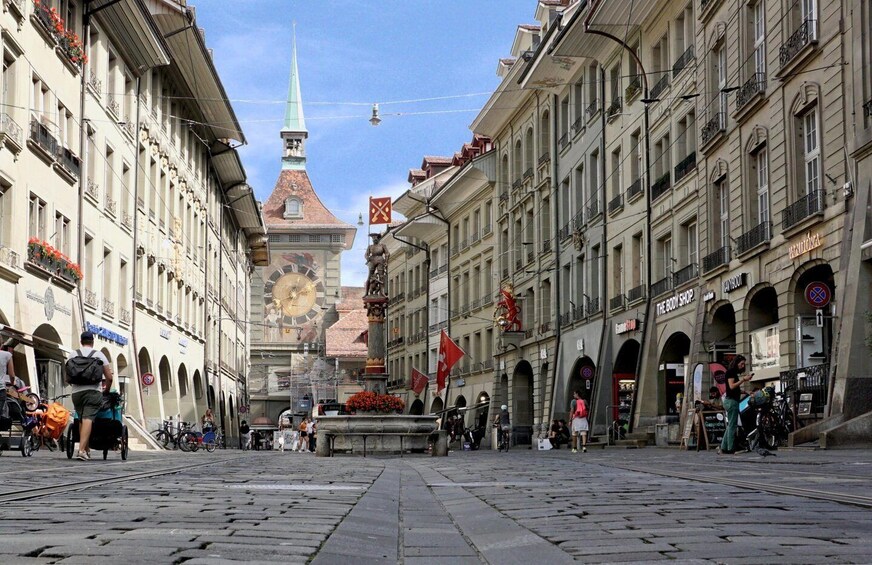 A cobblestone street with historic buildings, a clock tower, and a fountain in the center. 1