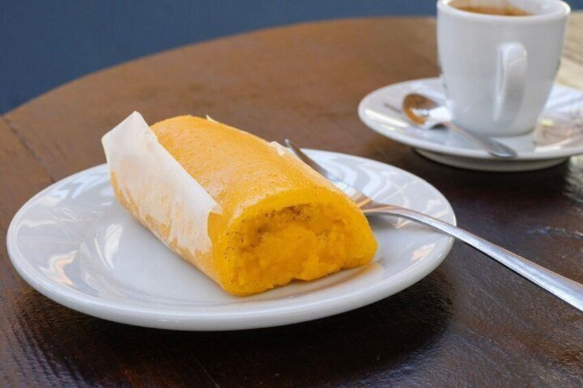 A rolled pastry on a white plate with a spoon, next to a cup of coffee on a saucer. 4