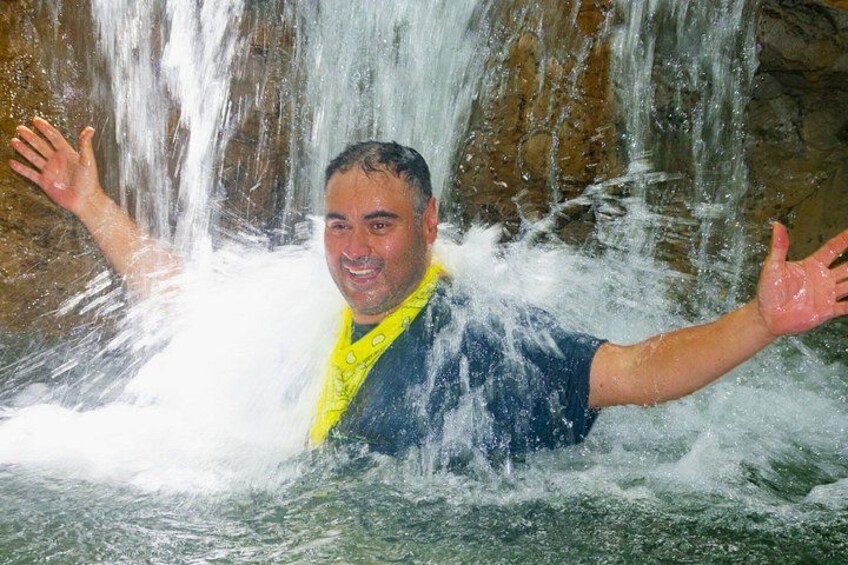A person in a blue shirt with arms outstretched, surrounded by water cascading from a rocky cliff. 2