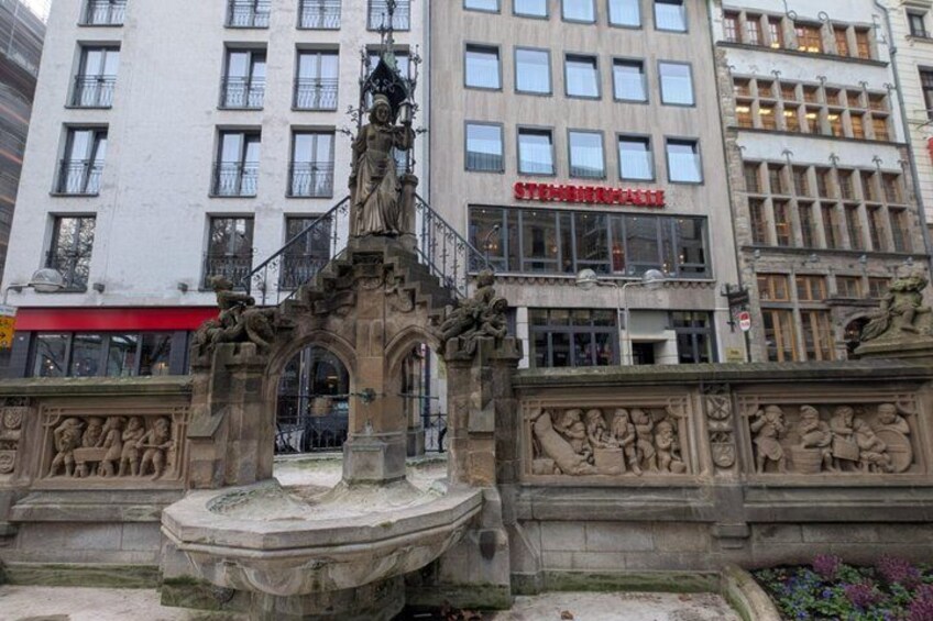 A historic fountain with intricate carvings, surrounded by a stone balustrade, in front of a modern building with a red awning. 3