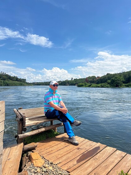 A man sitting on a wooden dock by a calm river, surrounded by greenery and a clear blue sky. 3