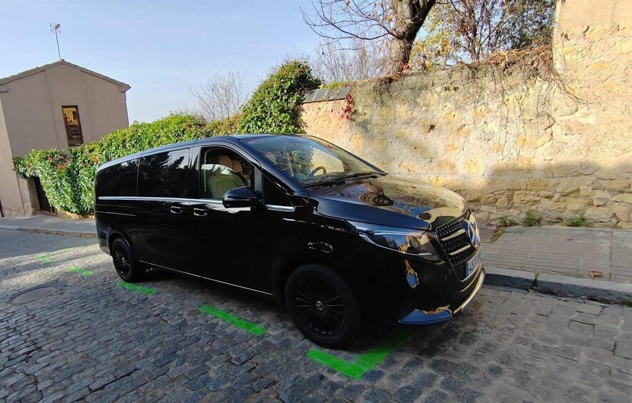 A black minivan parked on a cobblestone street with a stone wall and greenery in the background. 1