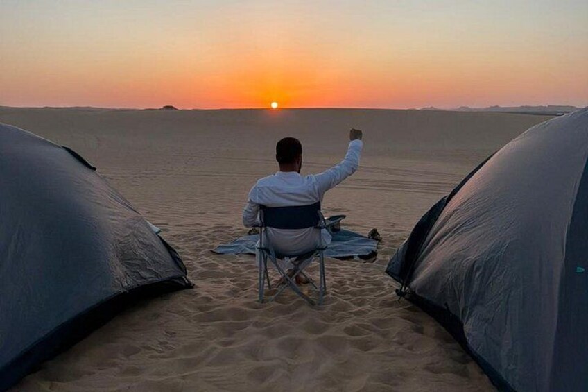 A person sitting on a chair facing a sunset over a desert with tents. 2