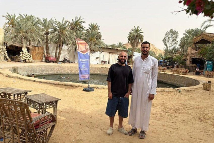 Two men standing in a sandy area with palm trees and a water feature in the background. 5