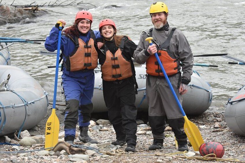 Three people wearing life jackets and helmets, standing near inflatable boats on a rocky shore. 5