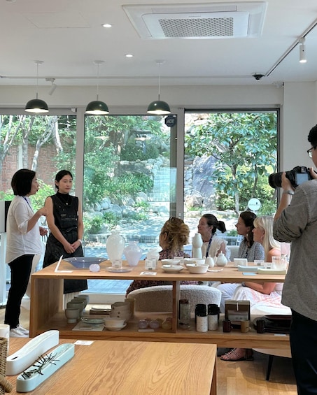 A group of people in a modern interior space with large windows, a table with ceramic items, and a person taking a photo. 5