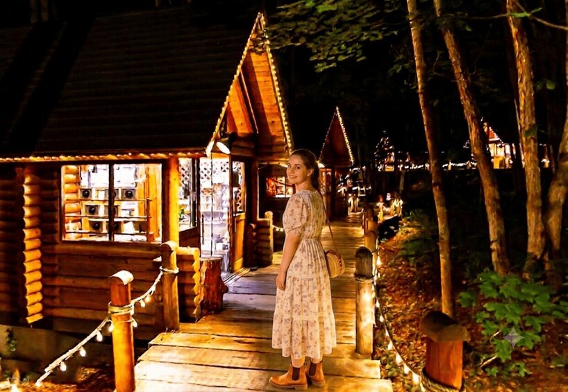 A wooden cabin with a porch, surrounded by trees and illuminated by string lights. 1