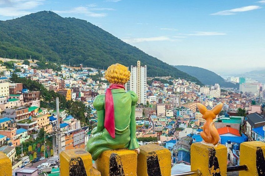A statue of a character with a yellow head and green body, sitting on a yellow barrier, overlooking a densely populated city with colorful buildings and a green hill in the background. 2