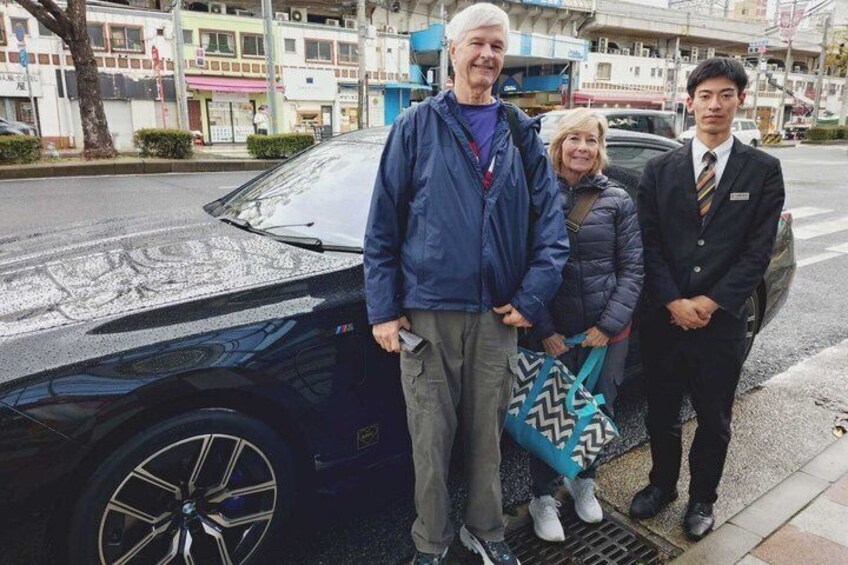 Three people standing in front of a black car on a city street. 1